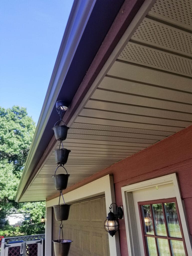 decorative-rain-chain-installation-roseville-mn A decorative dark metal cup-style rain chain installed on a dark gutter system of a red residential home in Roseville.