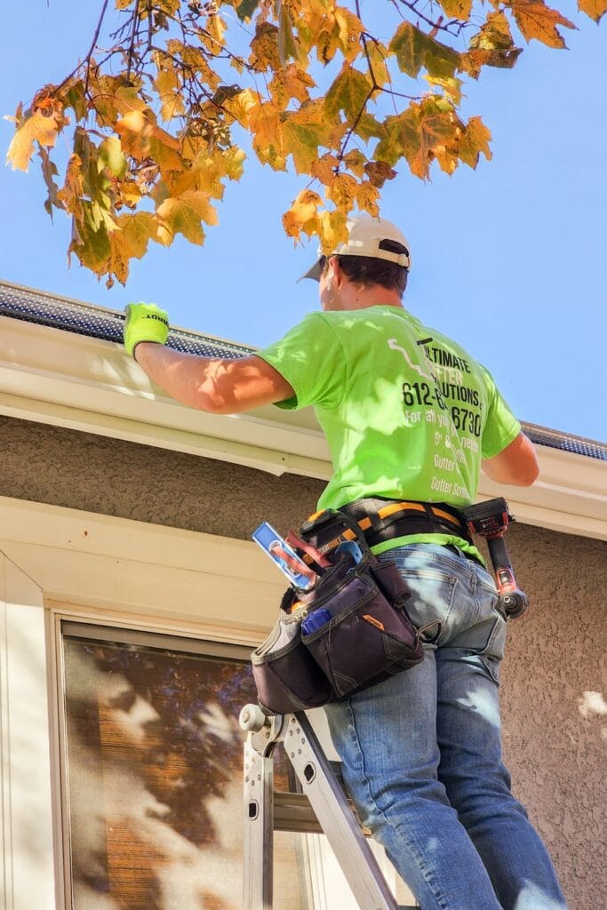 An Ultimate Gutter Solutions technician on a ladder installing a micro-mesh gutter guard under fall foliage in Plymouth.