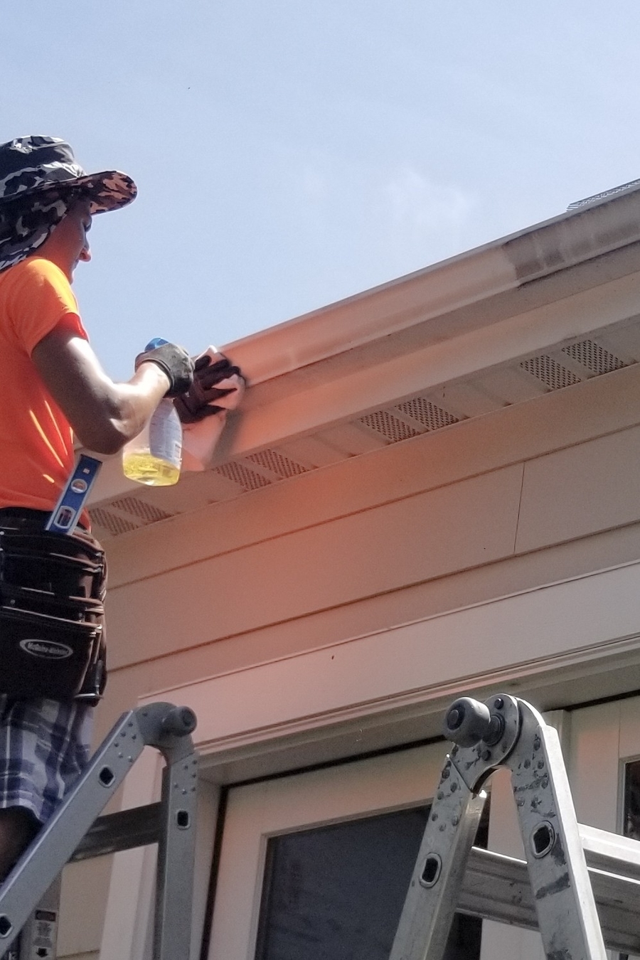 A professional cleaner on a ladder using a spray bottle and cloth to detail and clean the exterior of a white seamless gutter.