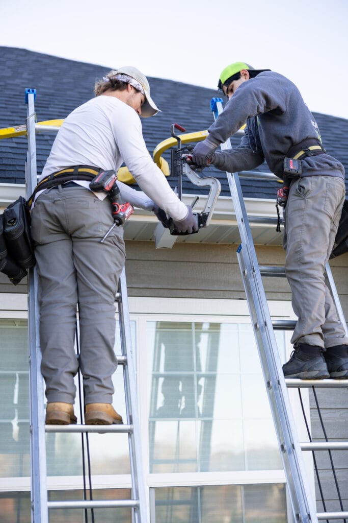Two professional contractors standing on ladders while installing a new gutter system on a residential home.