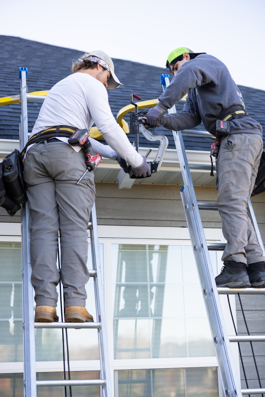 Two professional contractors standing on ladders while installing a new gutter system on a residential home.