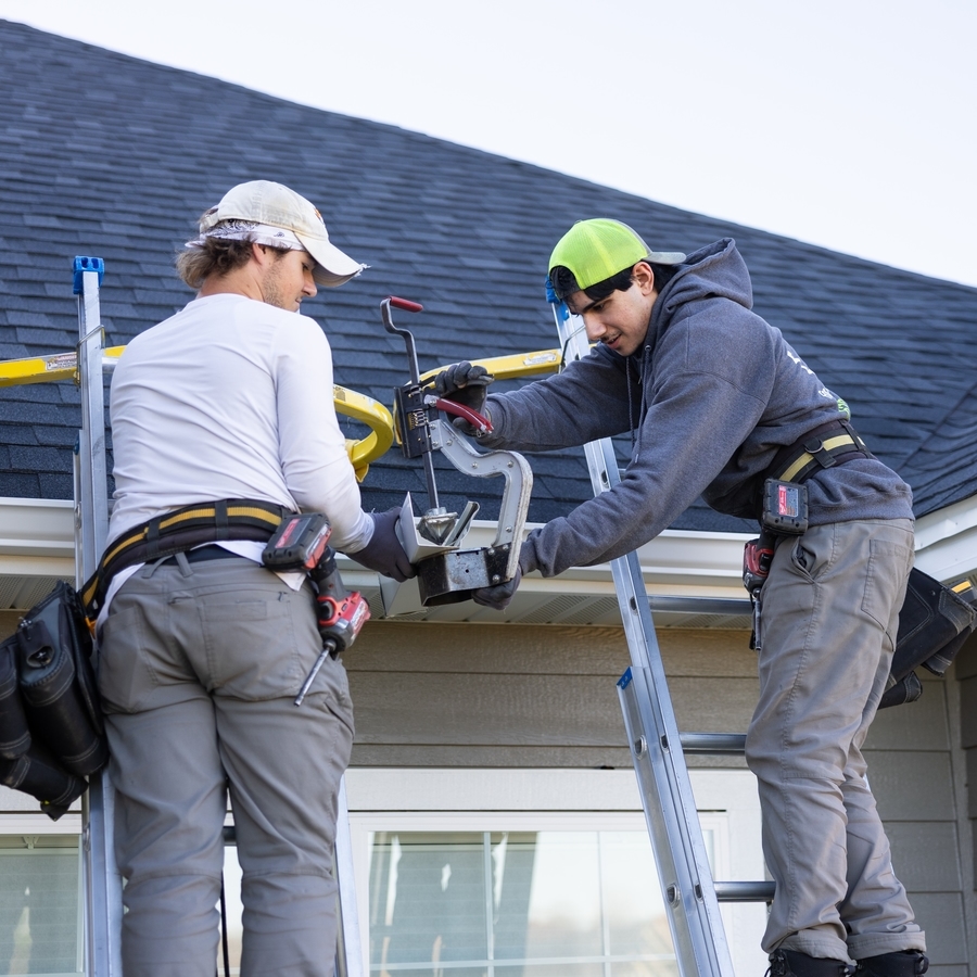 Two technicians on ladders using a professional gutter miter tool to join white gutter sections on a residential roof.